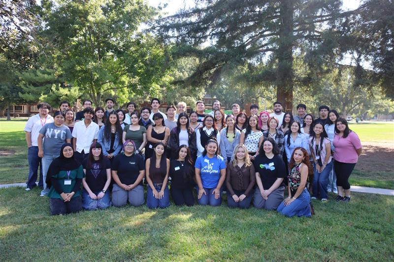 Group photo of about 50 people seated and standing on grass in a tree-shaded park