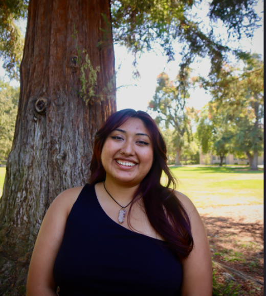 Smiling young woman in a black one-shoulder top standing by a large tree in a sunlit park