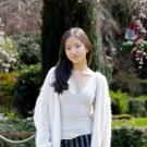 A young woman in a white cardigan stands in a lush garden with blooming flowers.